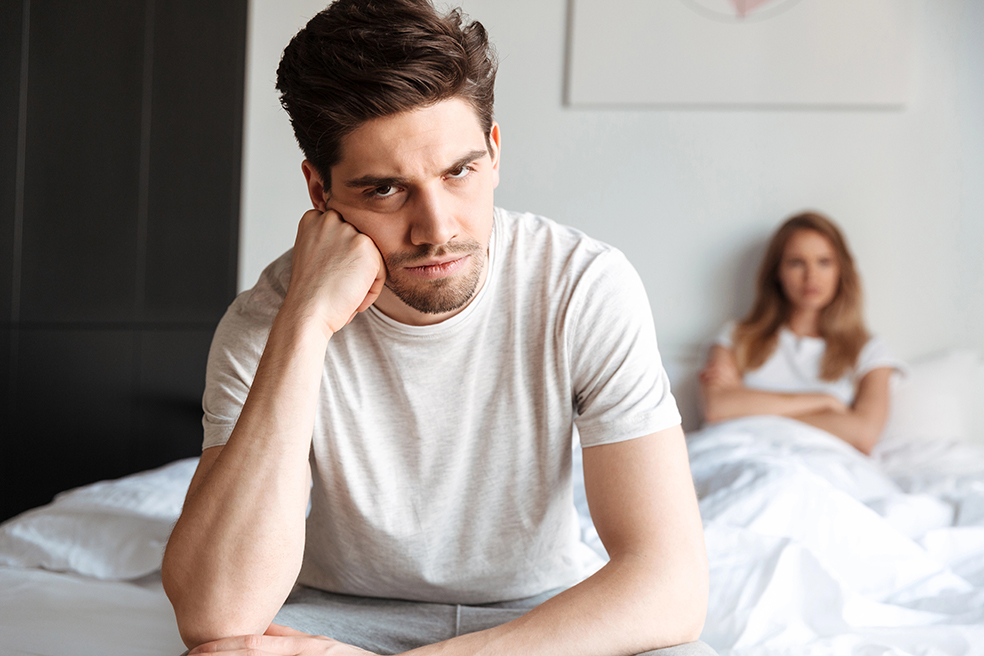 Upset man sitting on bed with angered wife in the background.